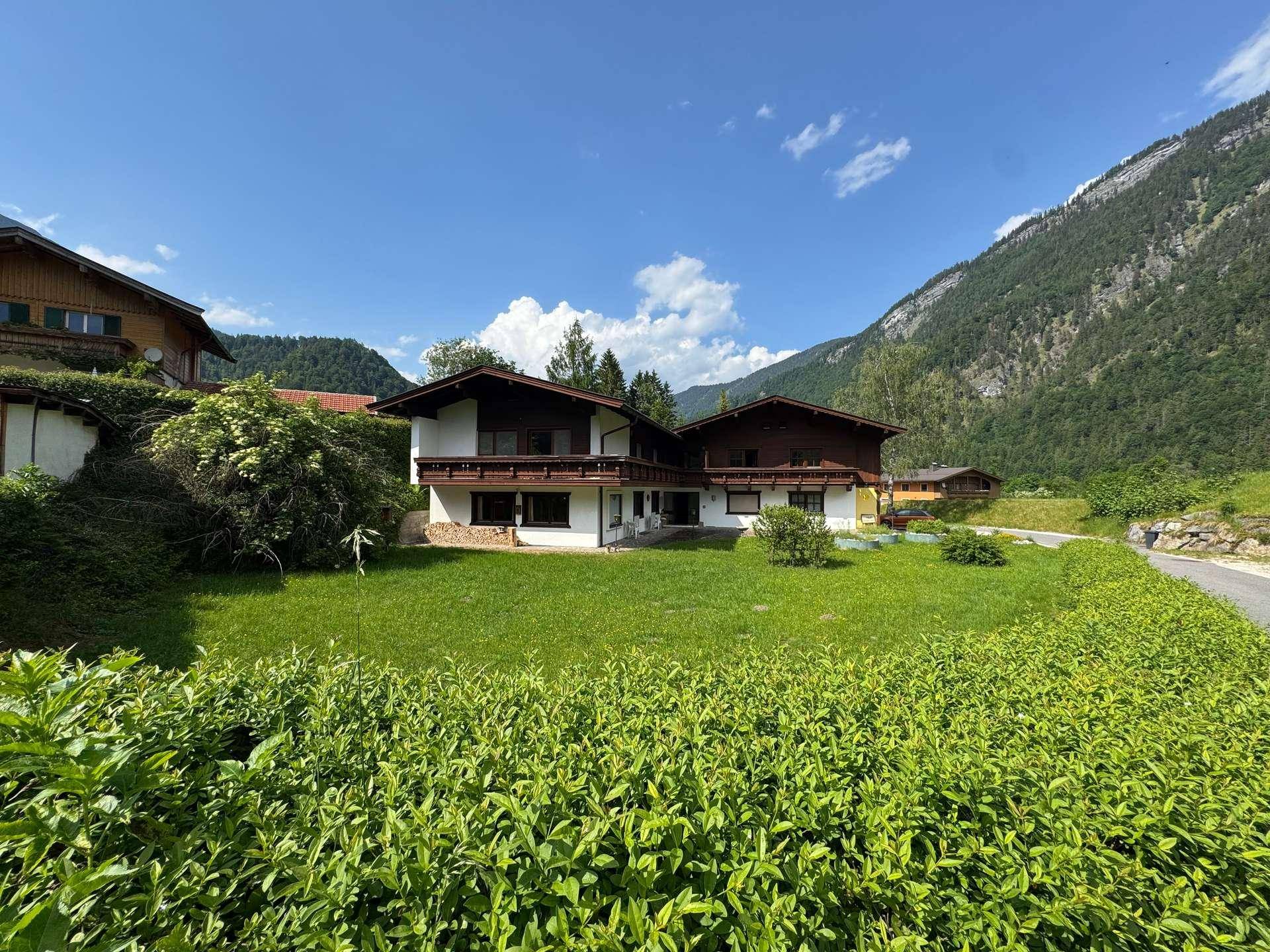 Haus in alpiner Landschaft mit großem Garten und Hecke, umgeben von Bergen.