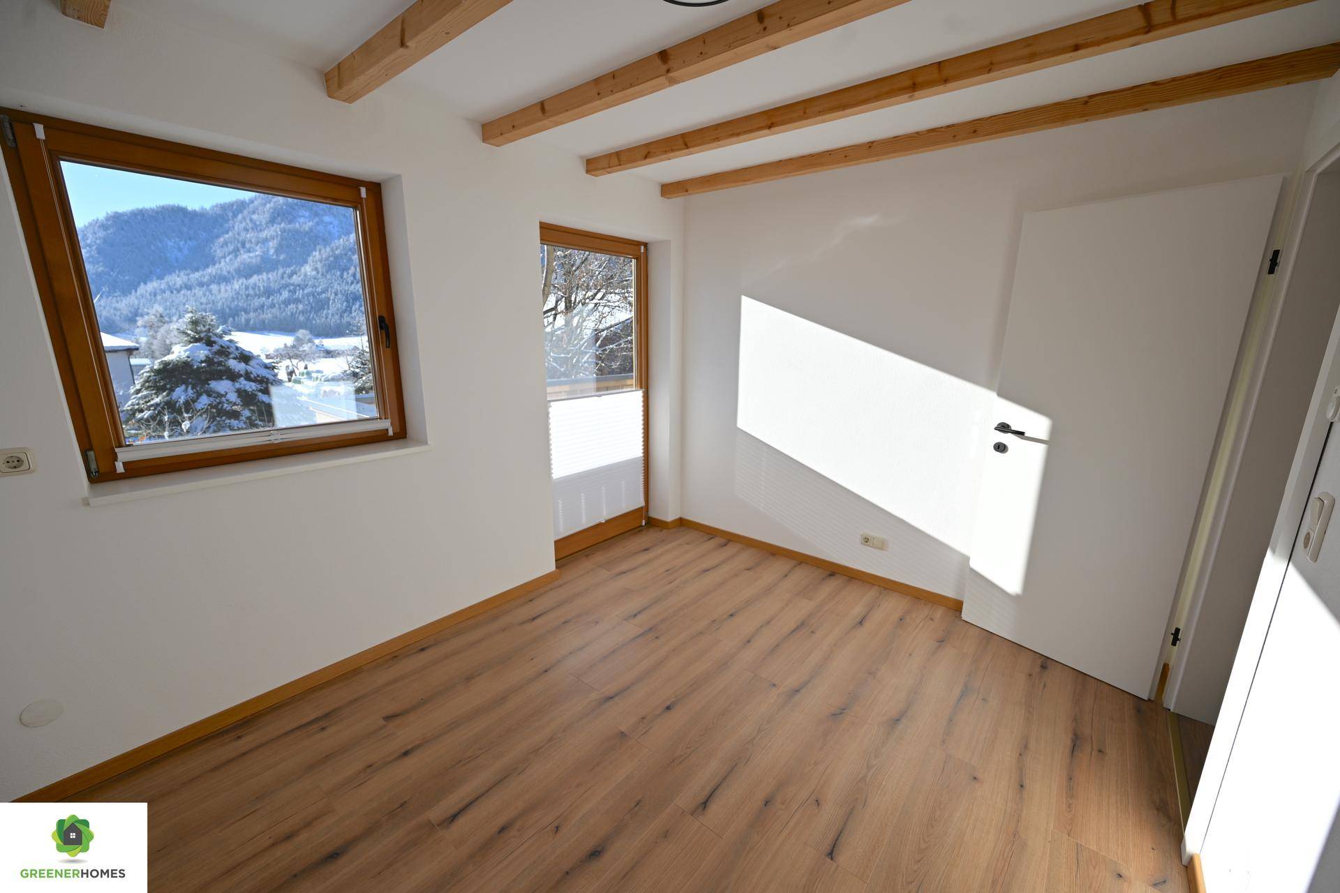 Helles Zimmer mit Laminatboden, Holzbalkendecke und einem Fenster mit Bergblick.
