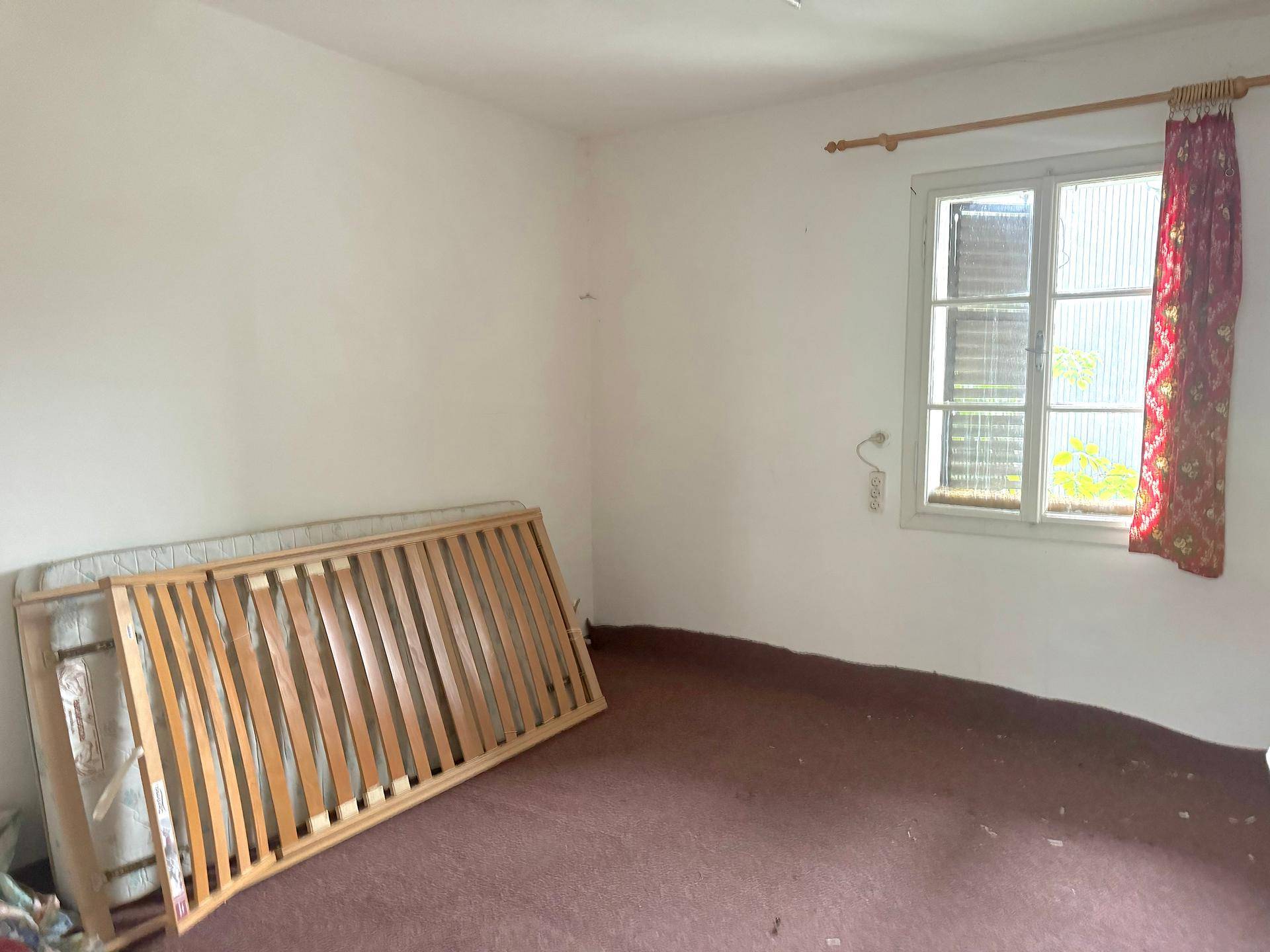 Schlafzimmer mit weiß gestrichenen Wänden, Teppichboden und einem Fenster mit rotem Vorhang.