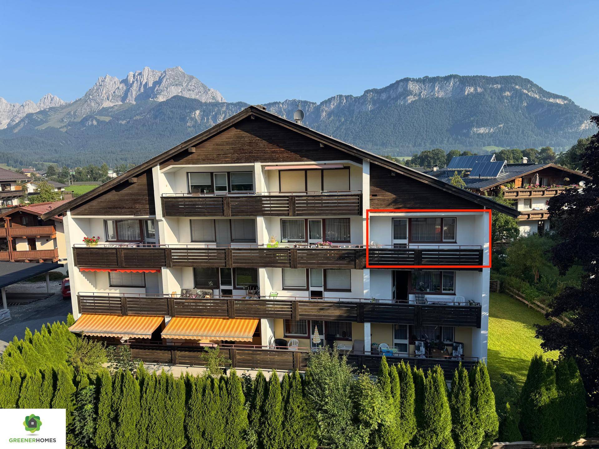 Mehrfamilienhaus mit Balkonen und Bergblick, umgeben von grüner Landschaft unter blauem Himmel.