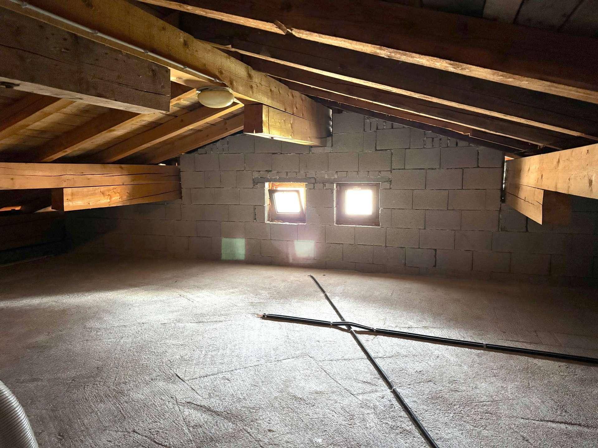Geräumiger Dachboden mit sichtbaren Holzbalken, Ziegelwänden und kleinen Fenstern.