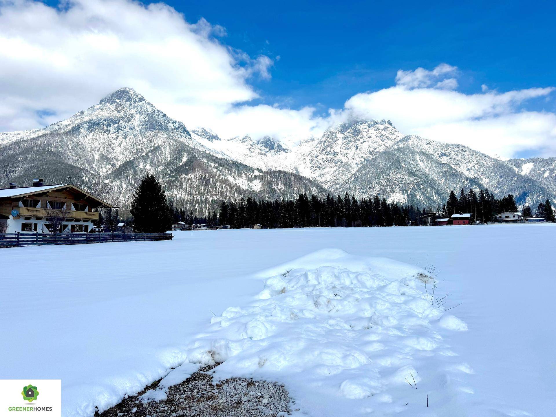 Winterliche Landschaft mit schneebedecktem Feld, traditionellen Häusern und majestätischen Bergen unter blauem Himmel.