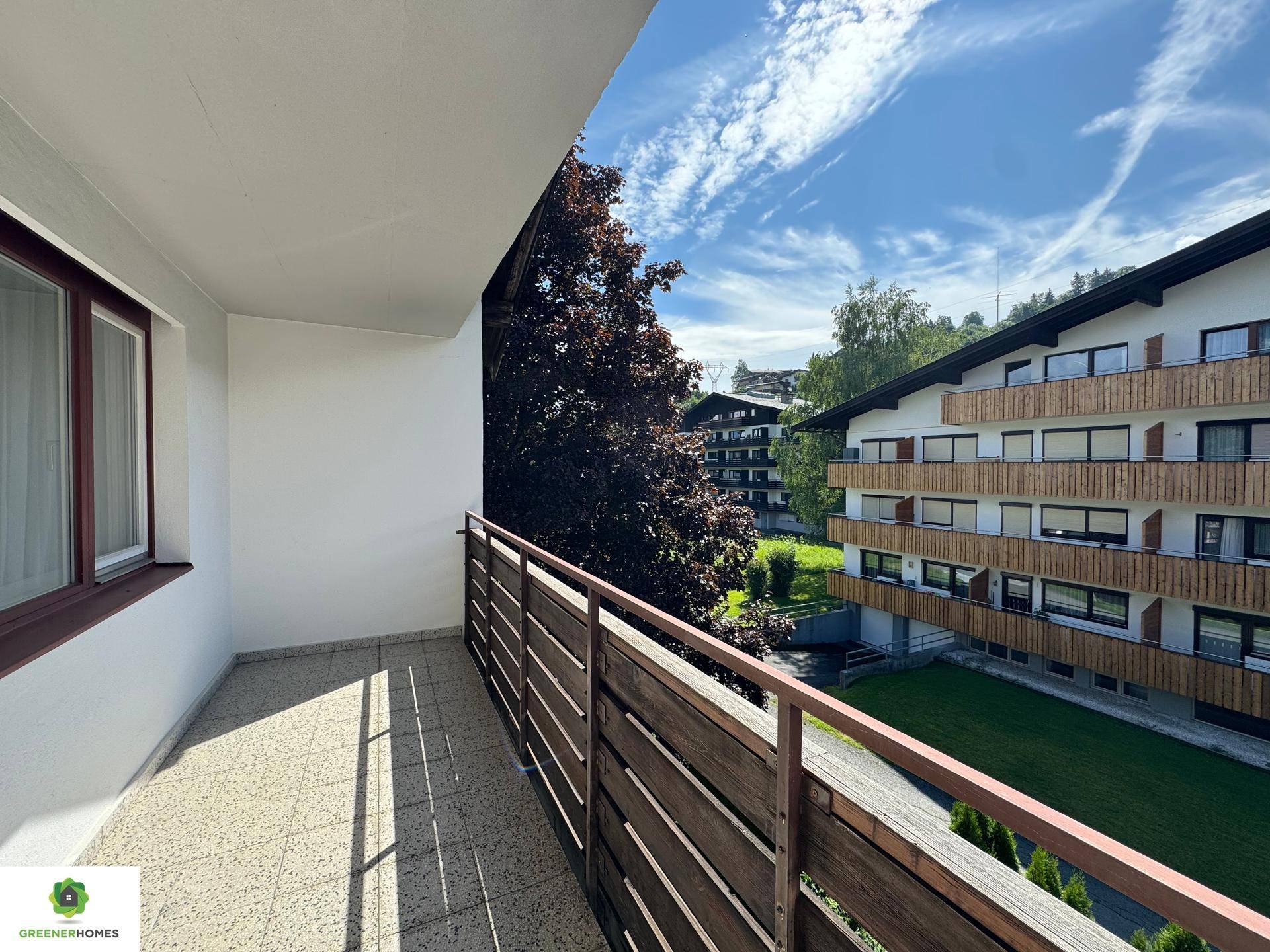 Geräumiger Balkon mit Fliesenboden und Holzgeländer, bietet Blick auf umliegende Gebäude und Grünflächen.