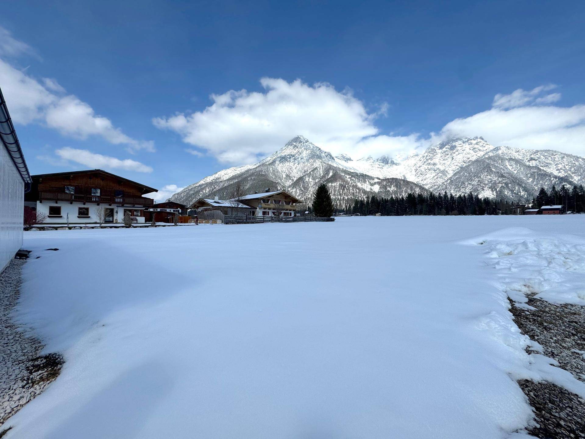Weitläufige Winterlandschaft mit tief verschneitem Feld, traditionellen Gebäuden und imposanter Bergkulisse.