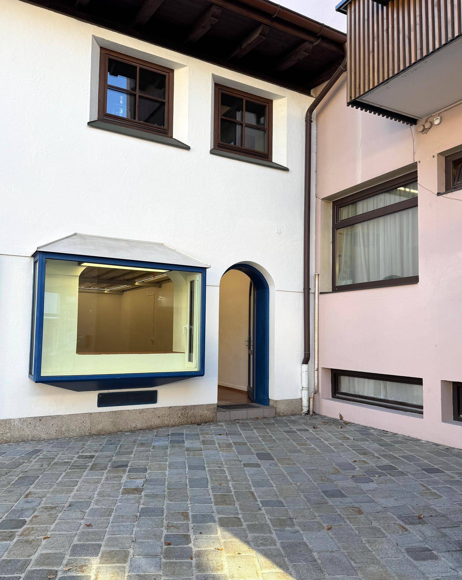 Gebäudefront mit gepflastertem Hof, einem blauen Schaufenster und traditionellen Fenstern, die viel Tageslicht bieten.