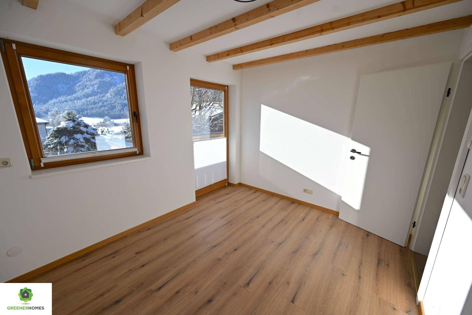 Leeres Zimmer mit Holzbalkendecke, Laminatboden und Blick auf verschneite Landschaft.