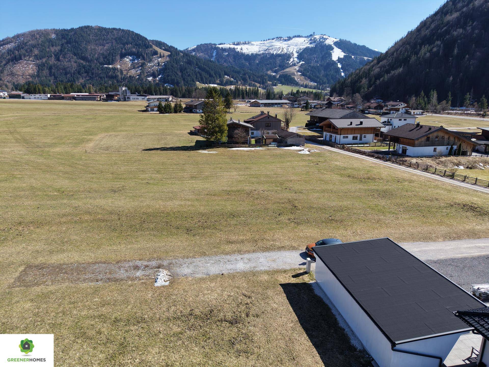 Luftaufnahme eines Wohngebiets mit Feldern und Häusern, umgeben von bewaldeten Hügeln und Bergen.