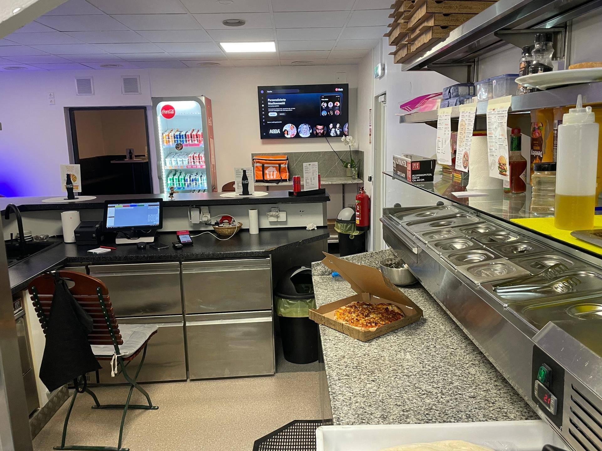 Professionelle Küche mit Edelstahloberflächen, Kühltheke und einem Bildschirm für Bestellungen.
