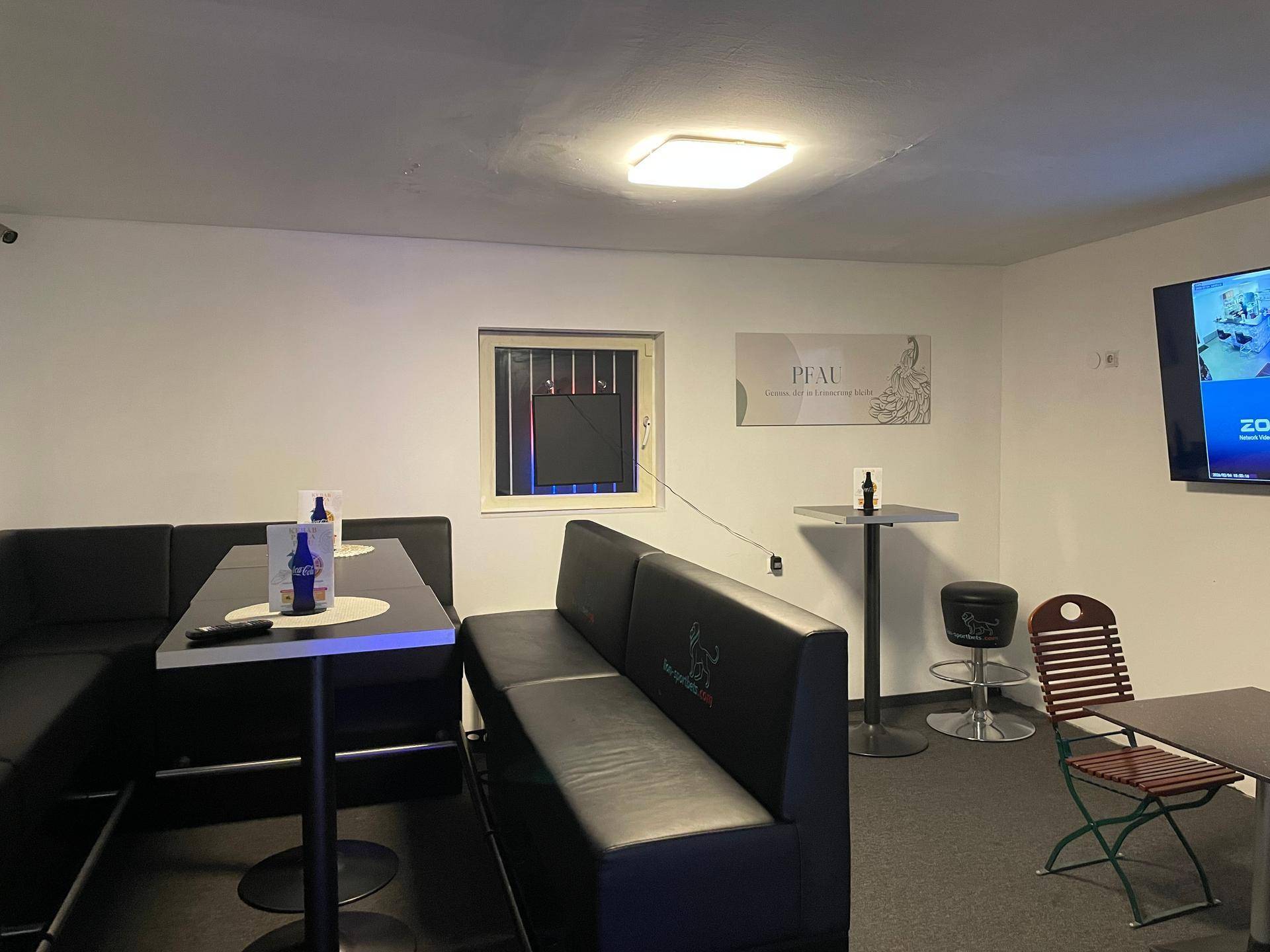 Loungebereich mit schwarzen Sitzbänken, Stehtischen und einem Fernseher an der Wand.