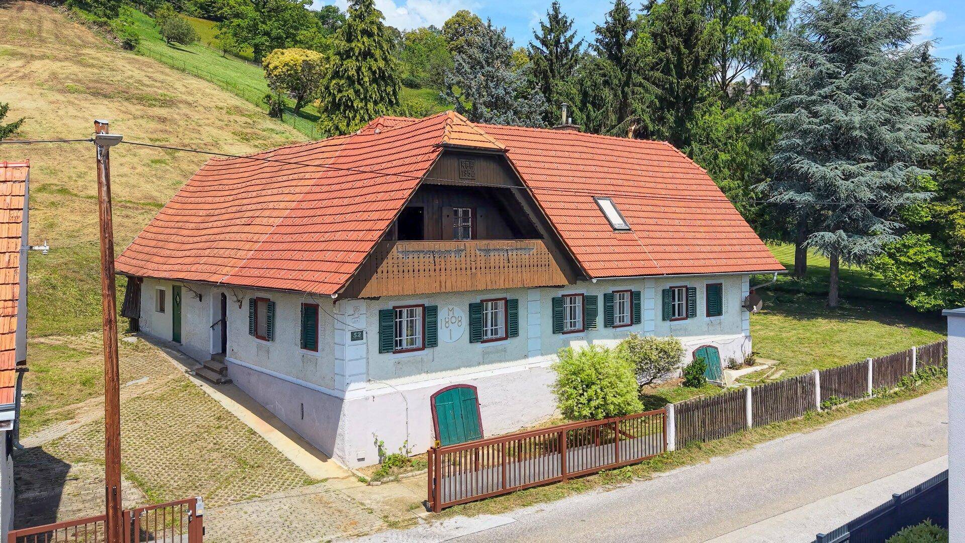 Traditionelles Wohnhaus mit rotem Ziegeldach, grünen Fensterläden und Holzbalkon an einer ländlichen Straße.