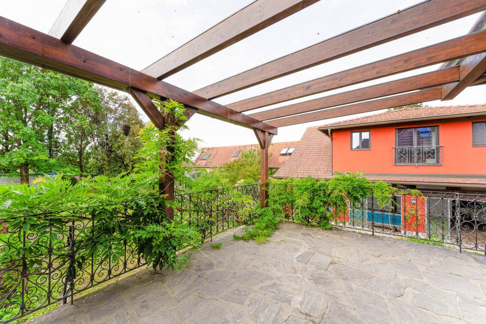 Große Steinterrasse mit Holzpergola und schmiedeeisernem Geländer, umgeben von Grünpflanzen.