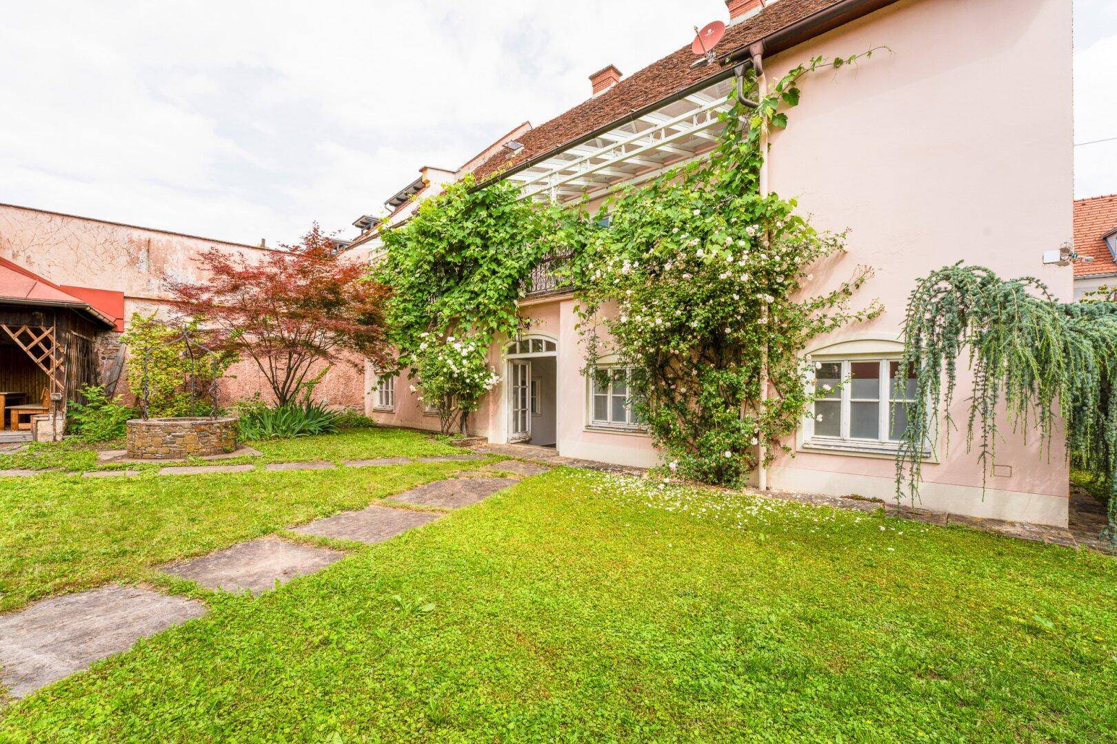 Grüner Gartenbereich vor einem hellrosa Gebäude mit Kletterpflanzen und einem überdachten Sitzplatz.