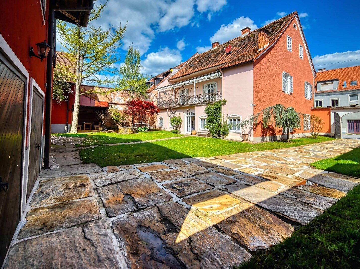Gepflasterter Hof mit Rasenfläche und einem hellrosa Gebäude mit Balkon im Hintergrund.