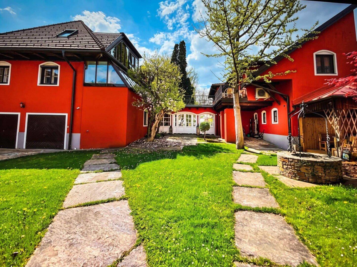 Rotes Haus mit gepflegtem Rasen und Steinwegen, umgeben von Bäumen und Sträuchern.