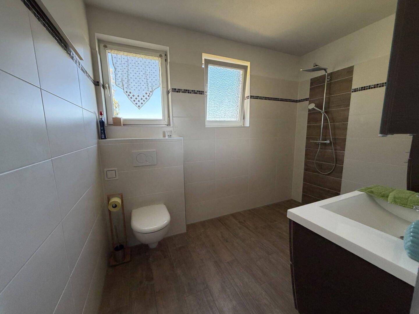 Modernes Badezimmer mit weißen Fliesen, Dusche und zwei Fenstern für Tageslicht.