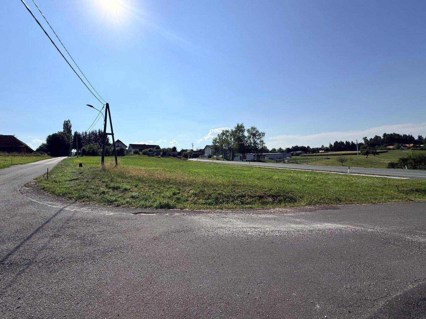 Grüne Wiese an einer Straßenkreuzung mit Häusern und Bäumen in der Ferne unter blauem Himmel.
