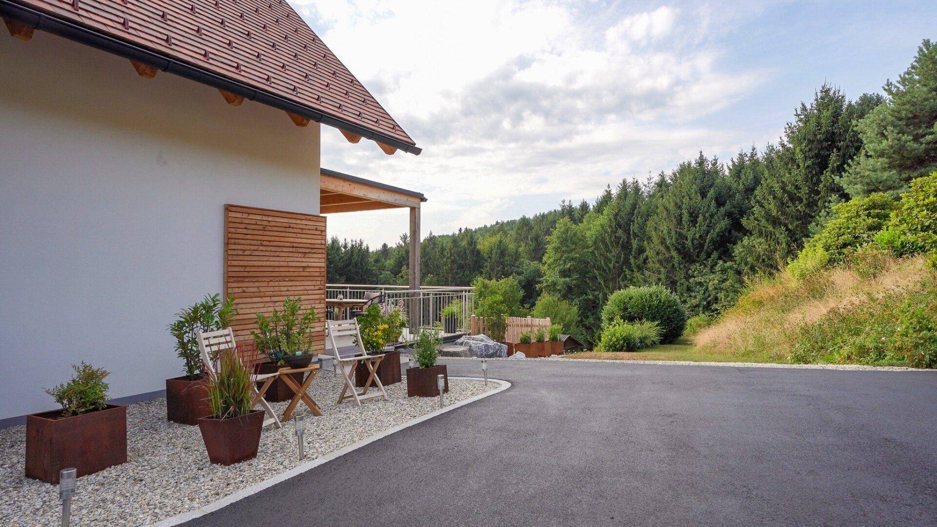 Zufahrt zum Haus mit Kiesflächen, bepflanzten Kübeln und Blick auf die bewaldete Umgebung.