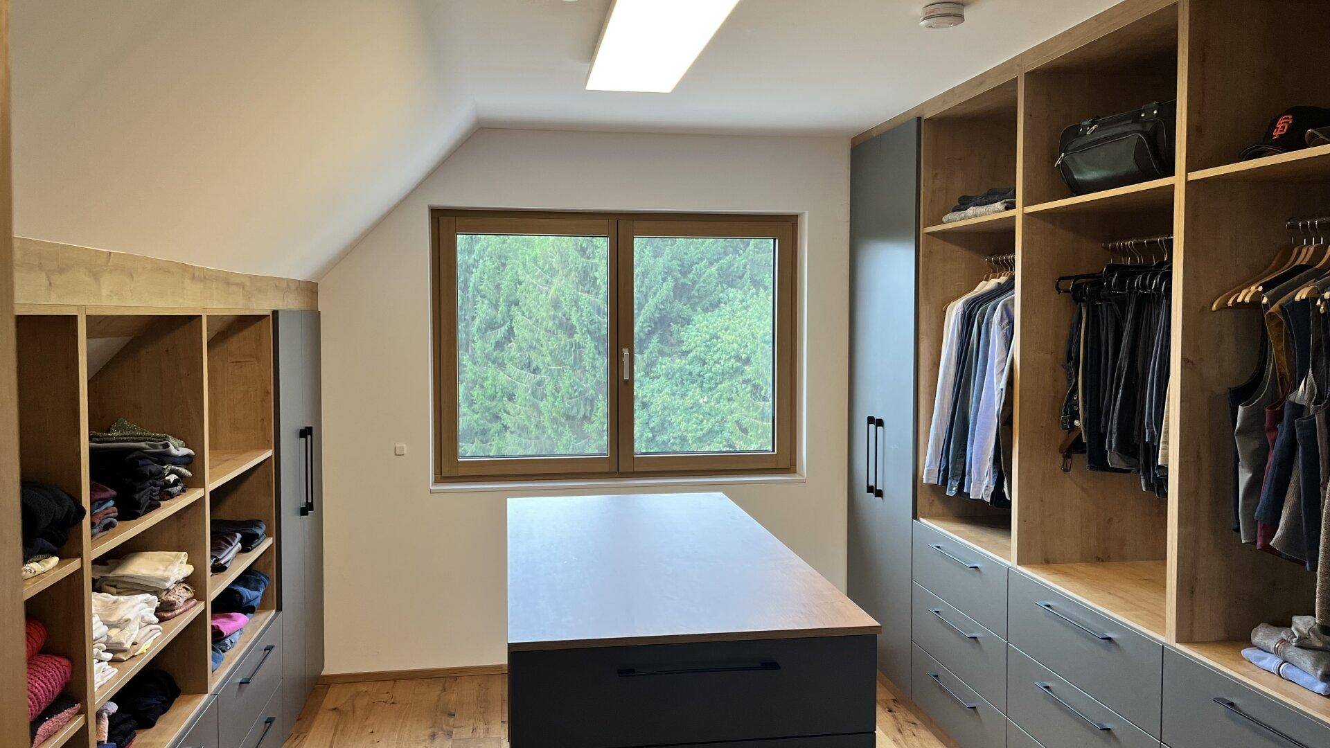 Großes Ankleidezimmer mit maßgefertigten Schränken, Mittelinsel und Fenster mit Blick ins Grüne.