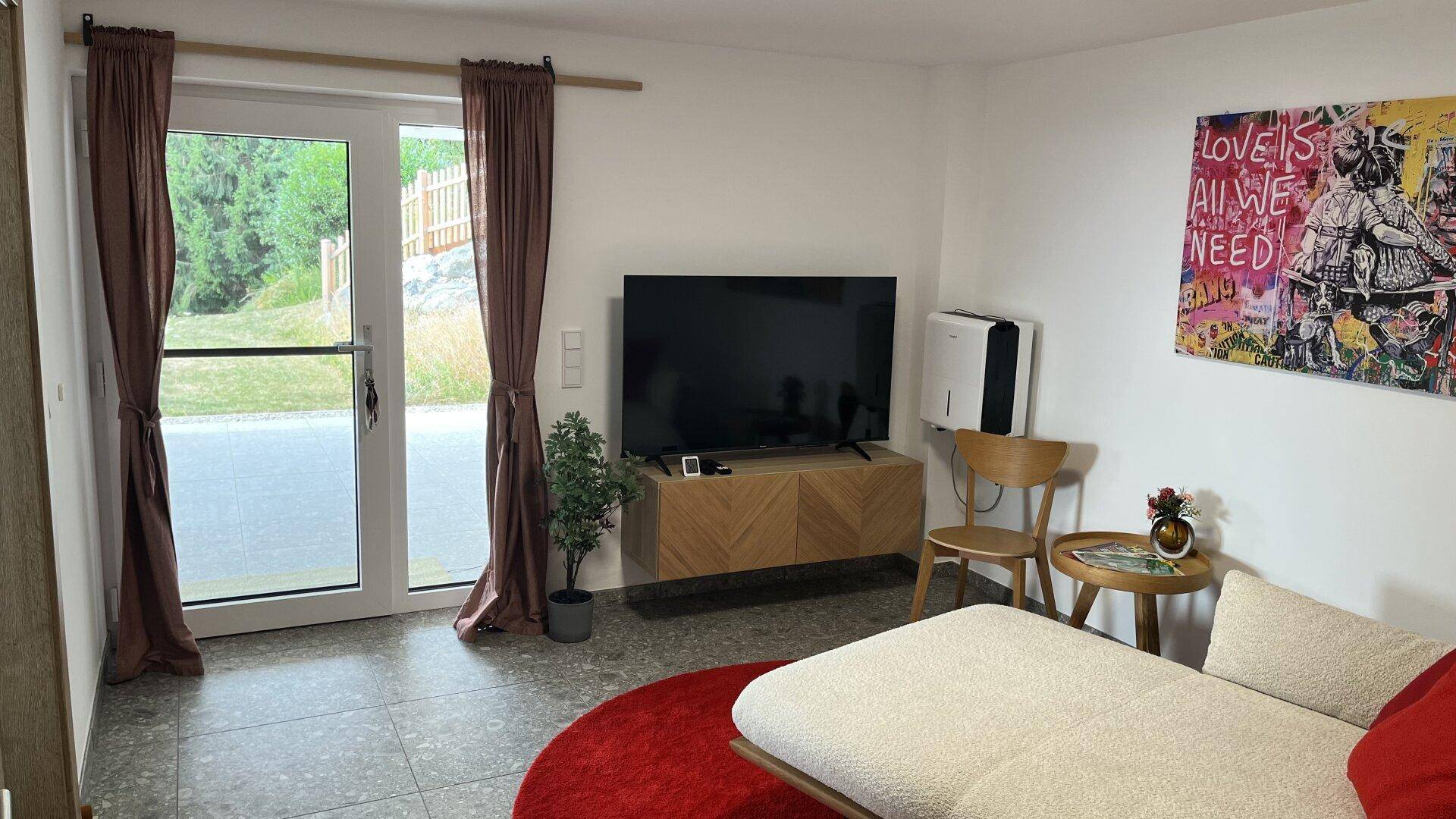 Helles Wohnzimmer mit Fliesenboden, Fernseher und direktem Zugang zur Terrasse.
