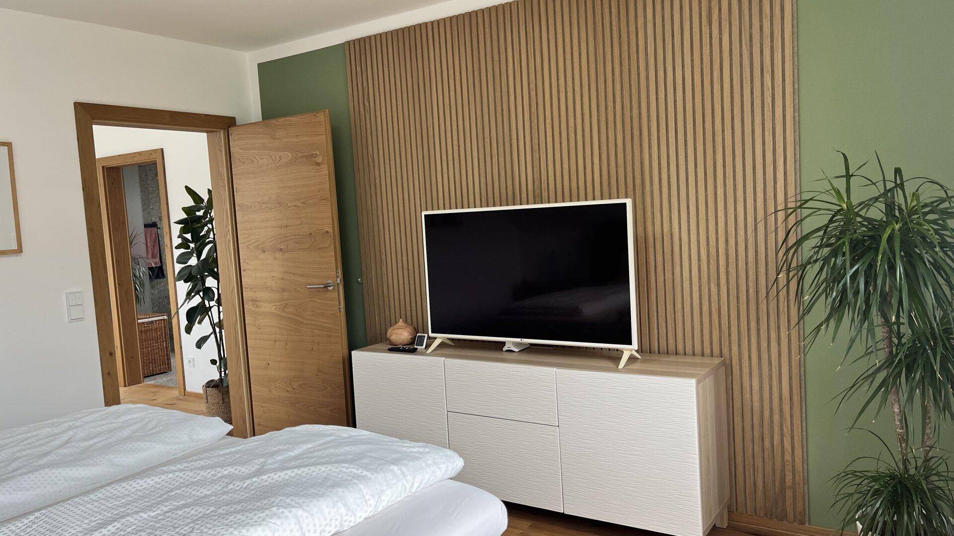 Schlafzimmer mit moderner Holzlamellenwand, Fernseher und Zugang zu einem weiteren Raum.