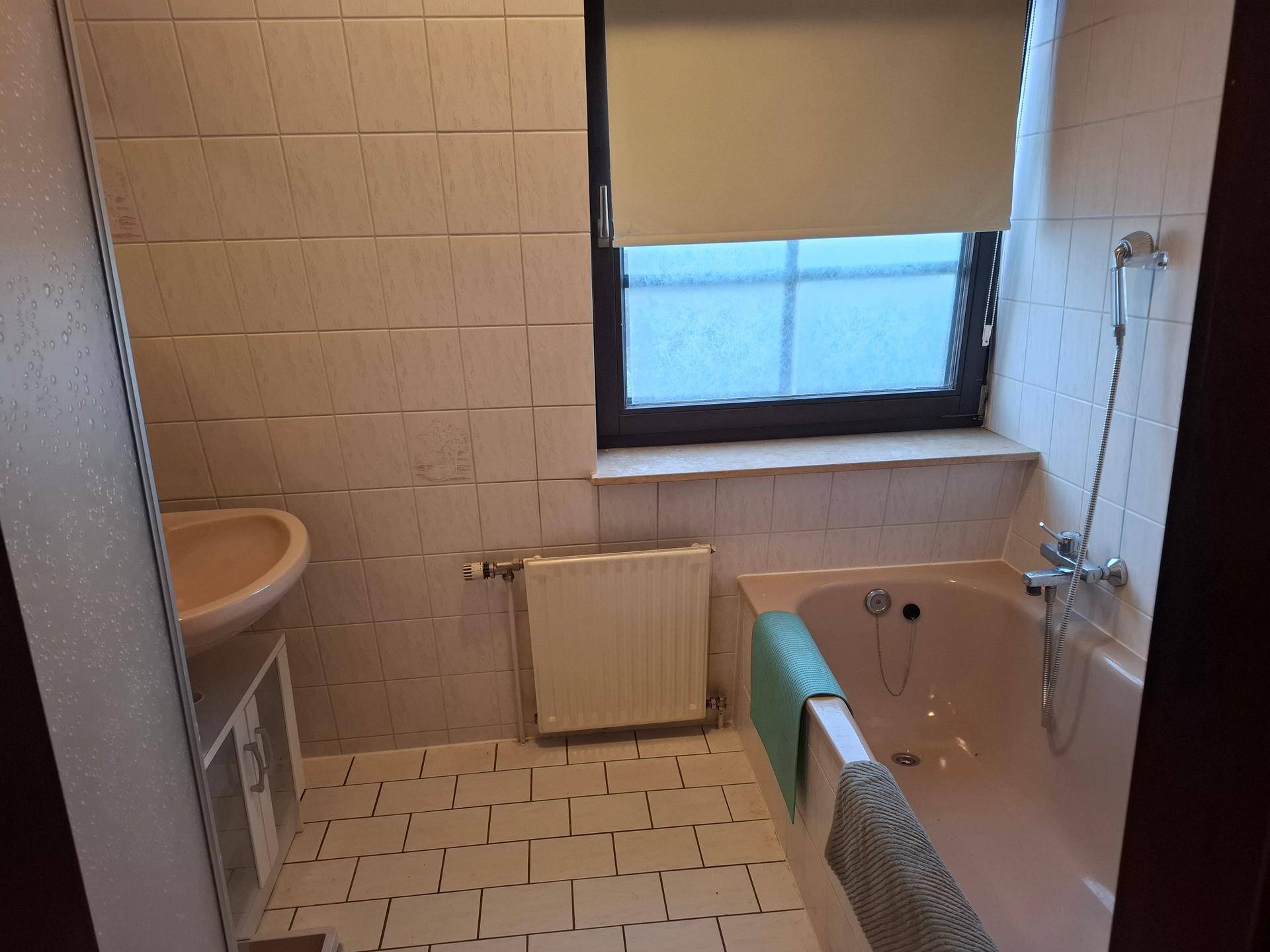 Badezimmer mit Badewanne, Waschbecken, Heizkörper und einem Fenster, alles in beigen Fliesen.
