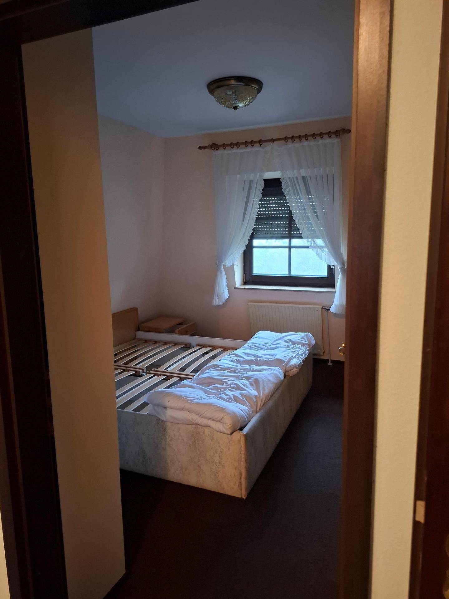 Schlafzimmer mit Bettgestell, dunklem Teppichboden und einem Fenster mit Rollladen und Vorhängen.