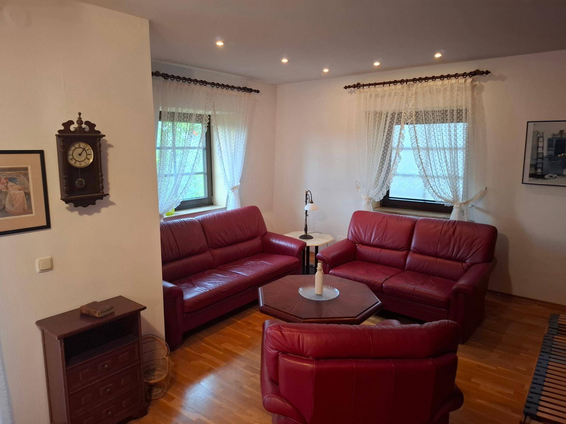 Wohnzimmer mit roten Ledersofas, Parkettboden und zwei Fenstern, die den Raum erhellen.