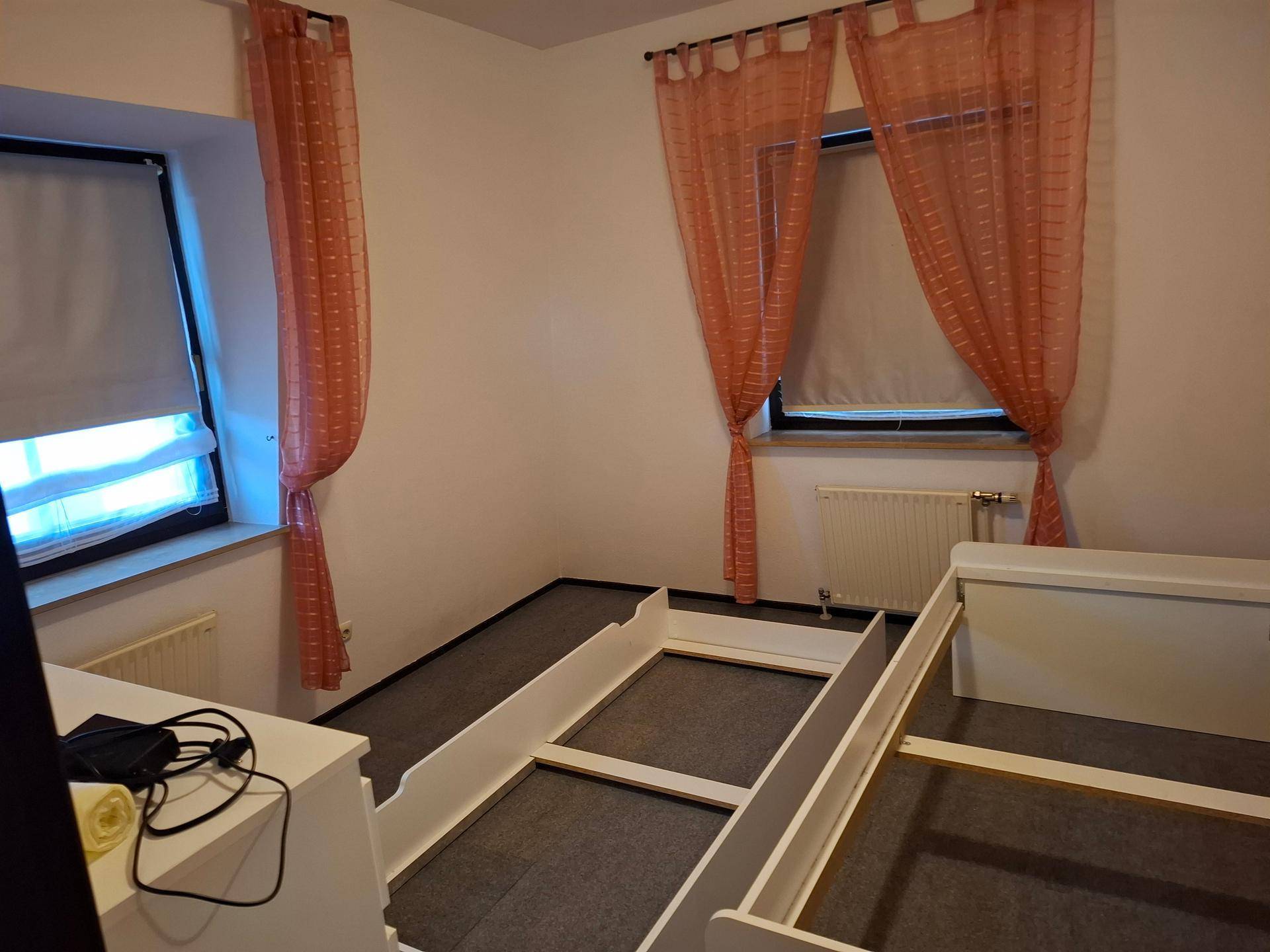 Schlafzimmer mit zwei Bettgestellen, grauem Teppichboden und Fenstern mit orangefarbenen Vorhängen.