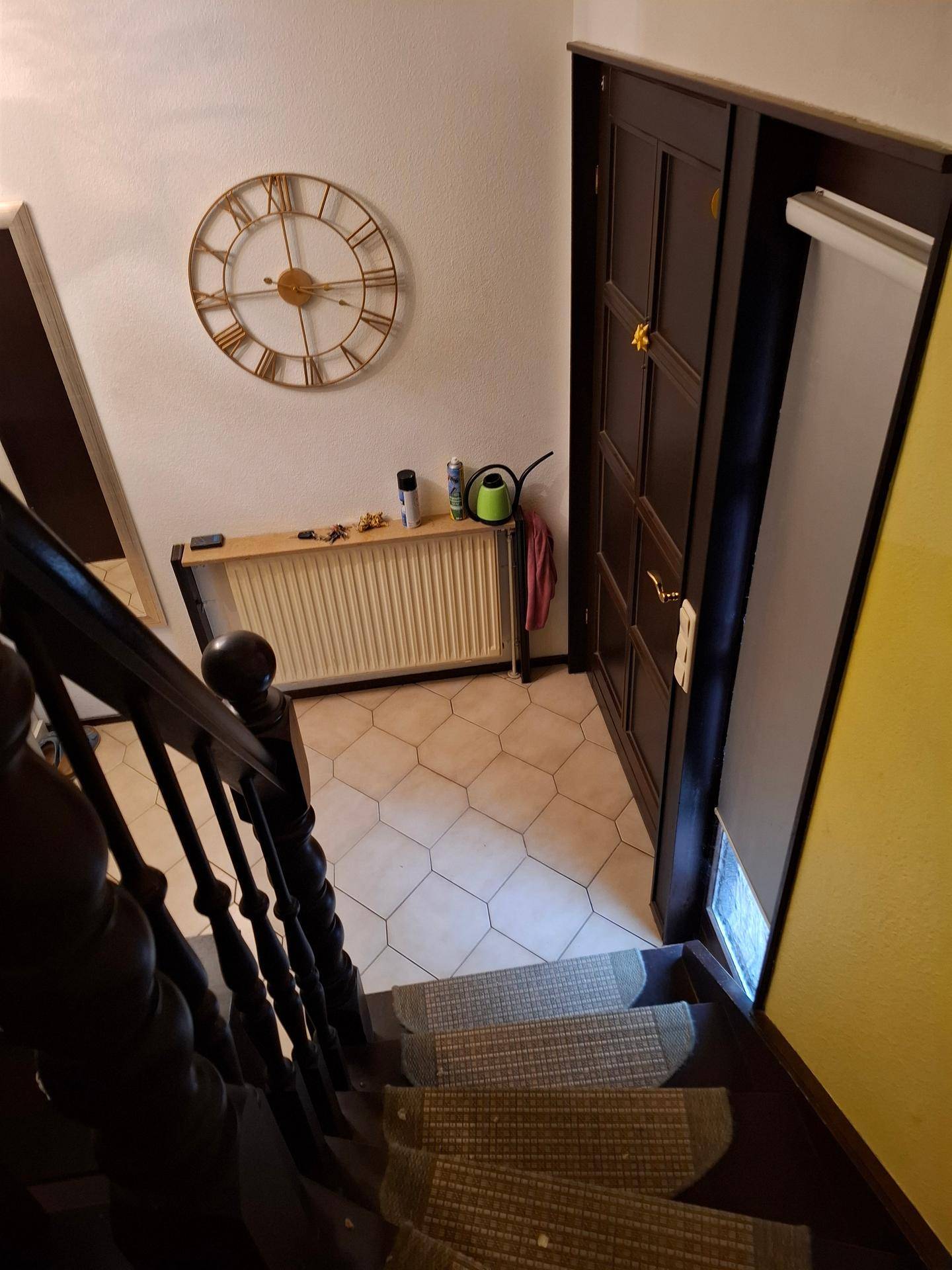 Blick von der Treppe in einen gefliesten Flur mit Heizkörper und einer großen Wanduhr.
