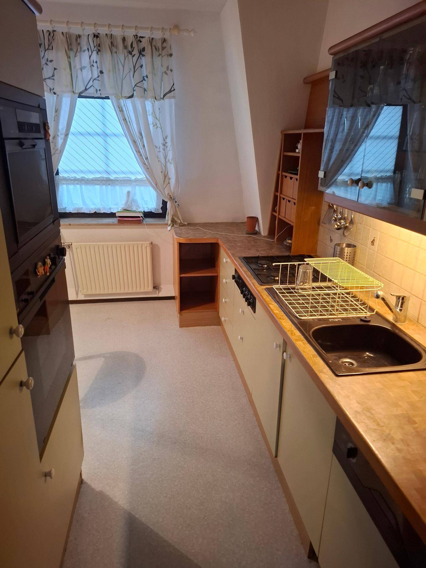 Küche im Dachgeschoss mit Schräge, älteren Geräten und einem Fenster mit Vorhängen.