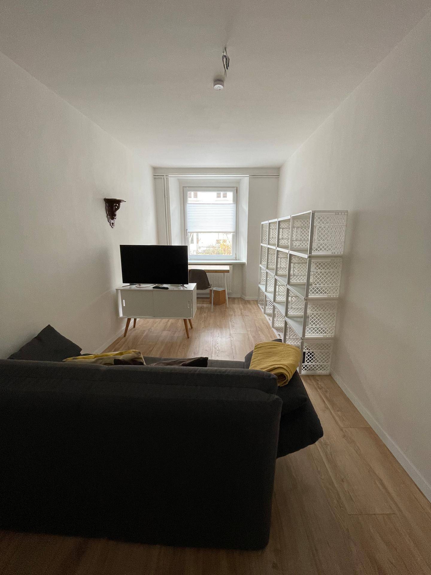 Schlichtes Wohnzimmer mit Sofa, Fernseher und hellem Holzfußboden, gut beleuchtet durch ein Fenster.