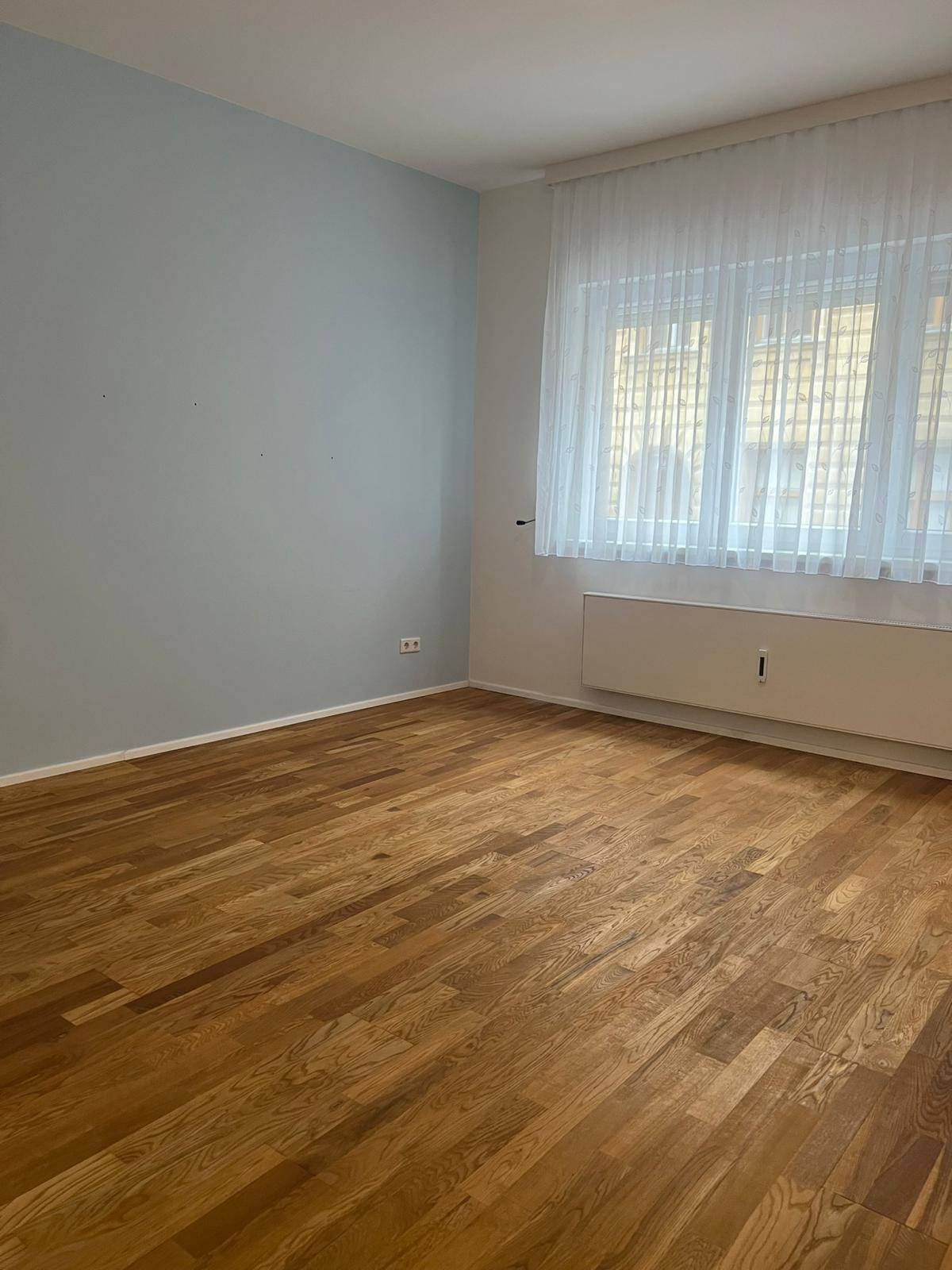 Helles Schlafzimmer mit Parkettboden, blauer Akzentwand und großem Fenster.