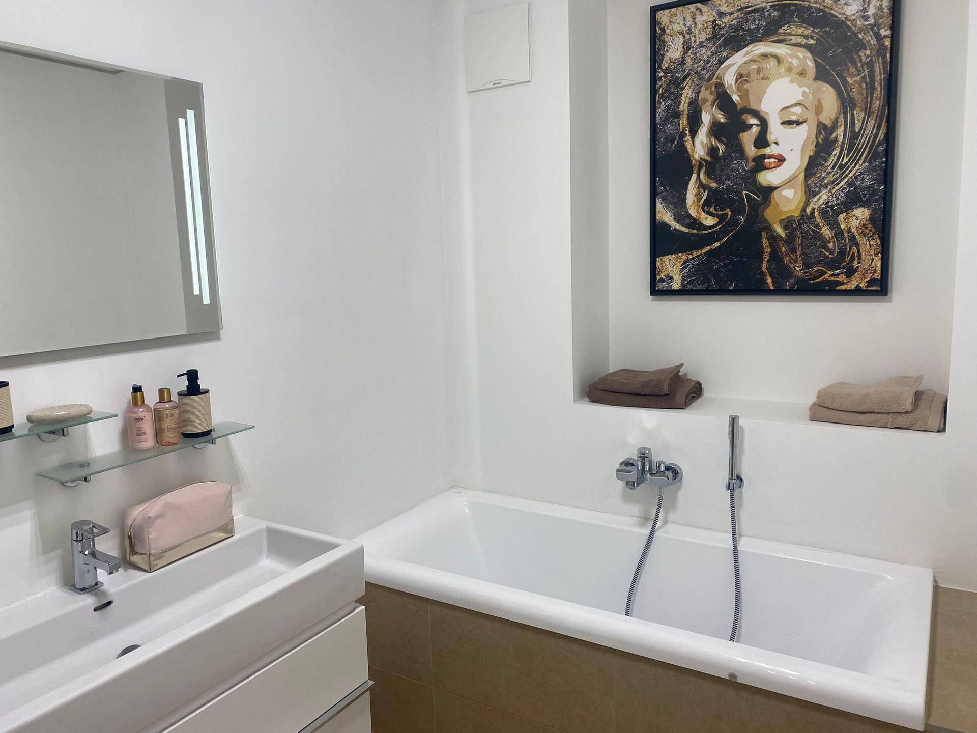 Helles Badezimmer mit integrierter Badewanne, Einzelwaschbecken und dekorativem Wandbild.