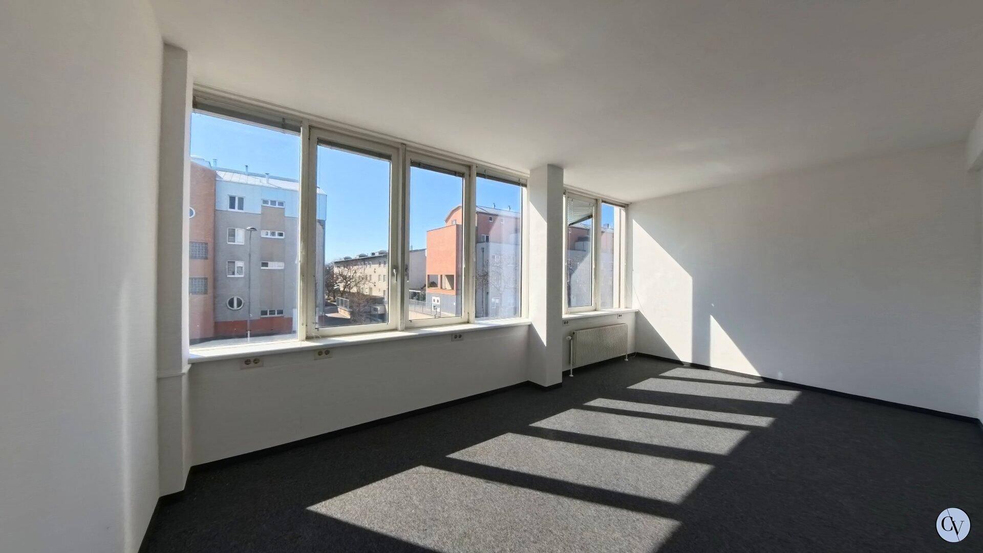 Heller Büroraum mit großen Fenstern, die viel Tageslicht hereinlassen und einen Blick auf umliegende Gebäude bieten.