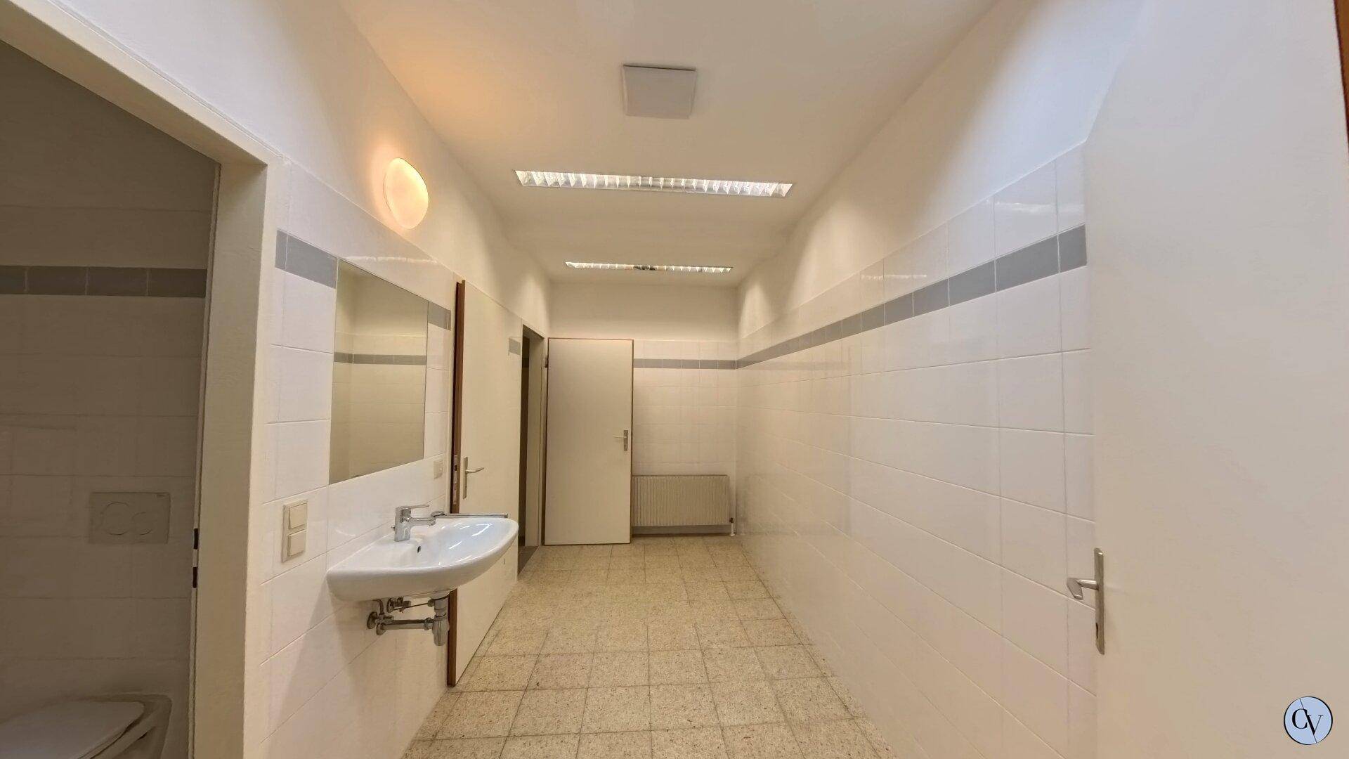Geräumiger, gefliester Sanitärbereich mit Waschbecken und Spiegel, der einen funktionalen Eindruck macht.