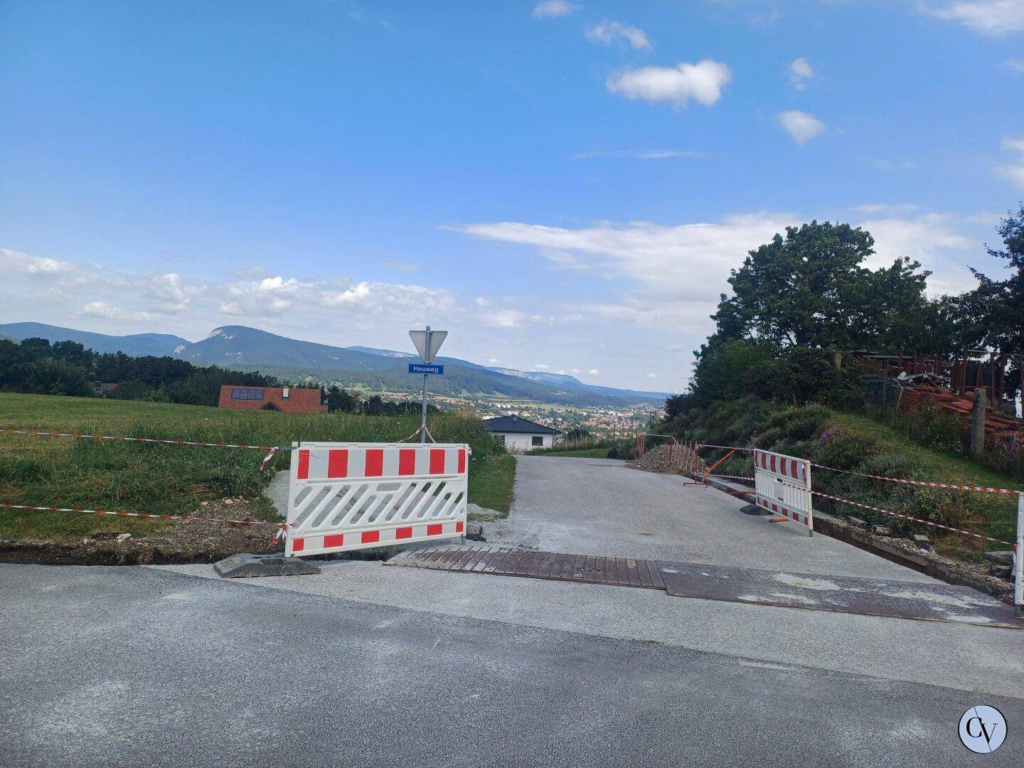 Blick über eine ländliche Straße mit Baustellenabsperrungen und weitreichender Aussicht auf eine Stadt und Berge.