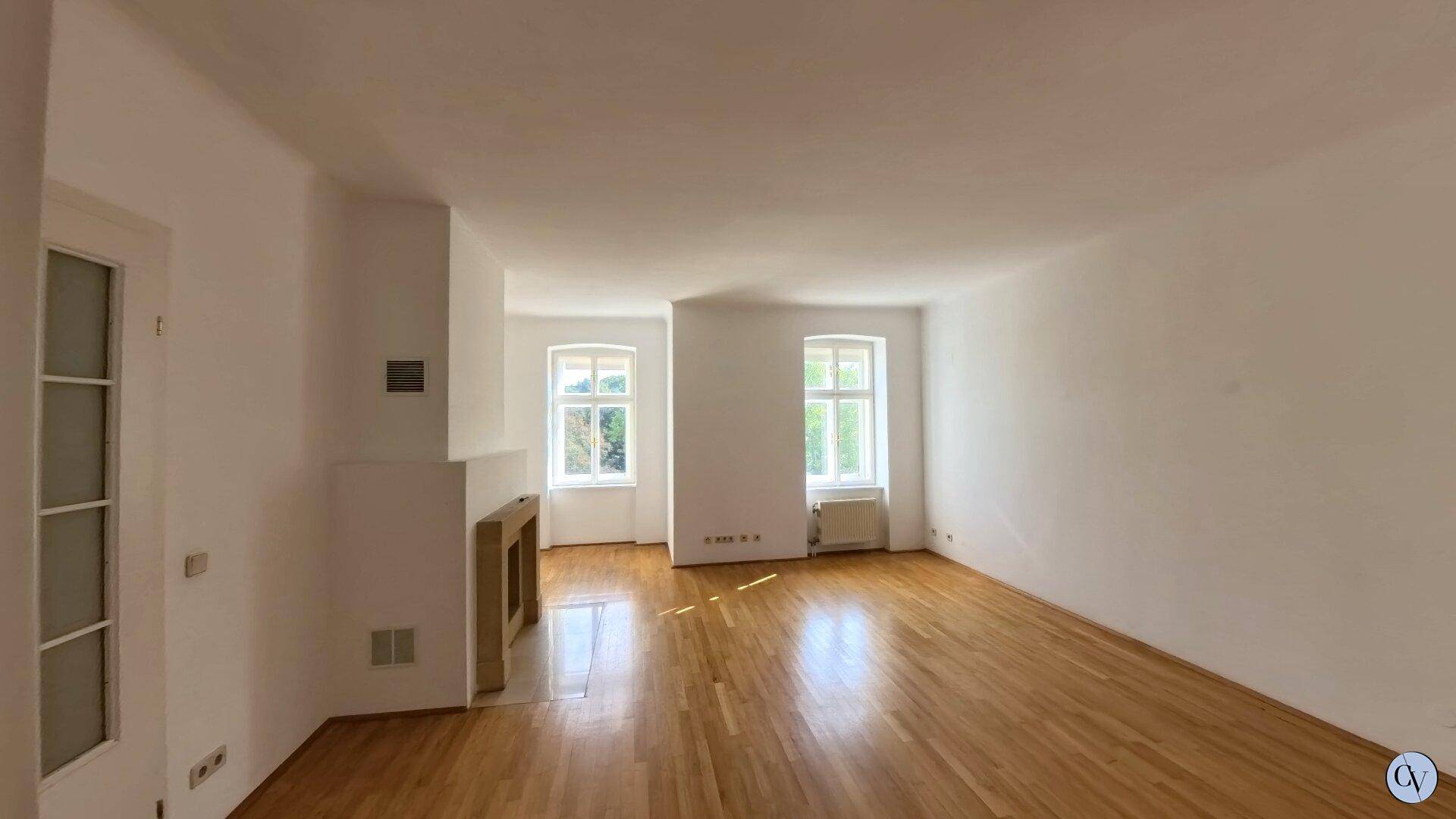 Geräumiges Wohnzimmer mit Parkettboden, Kamin und zwei klassischen Fenstern.