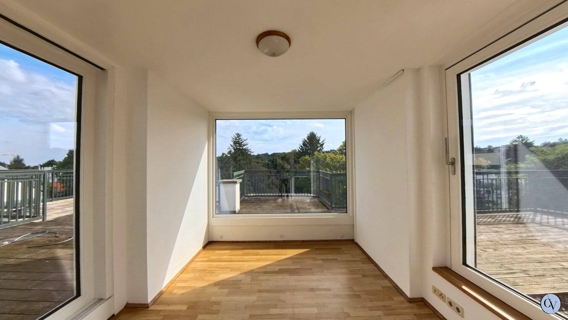 Heller Raum mit großen Fenstern, Holzparkett und weitem Blick ins Grüne sowie auf eine Terrasse.