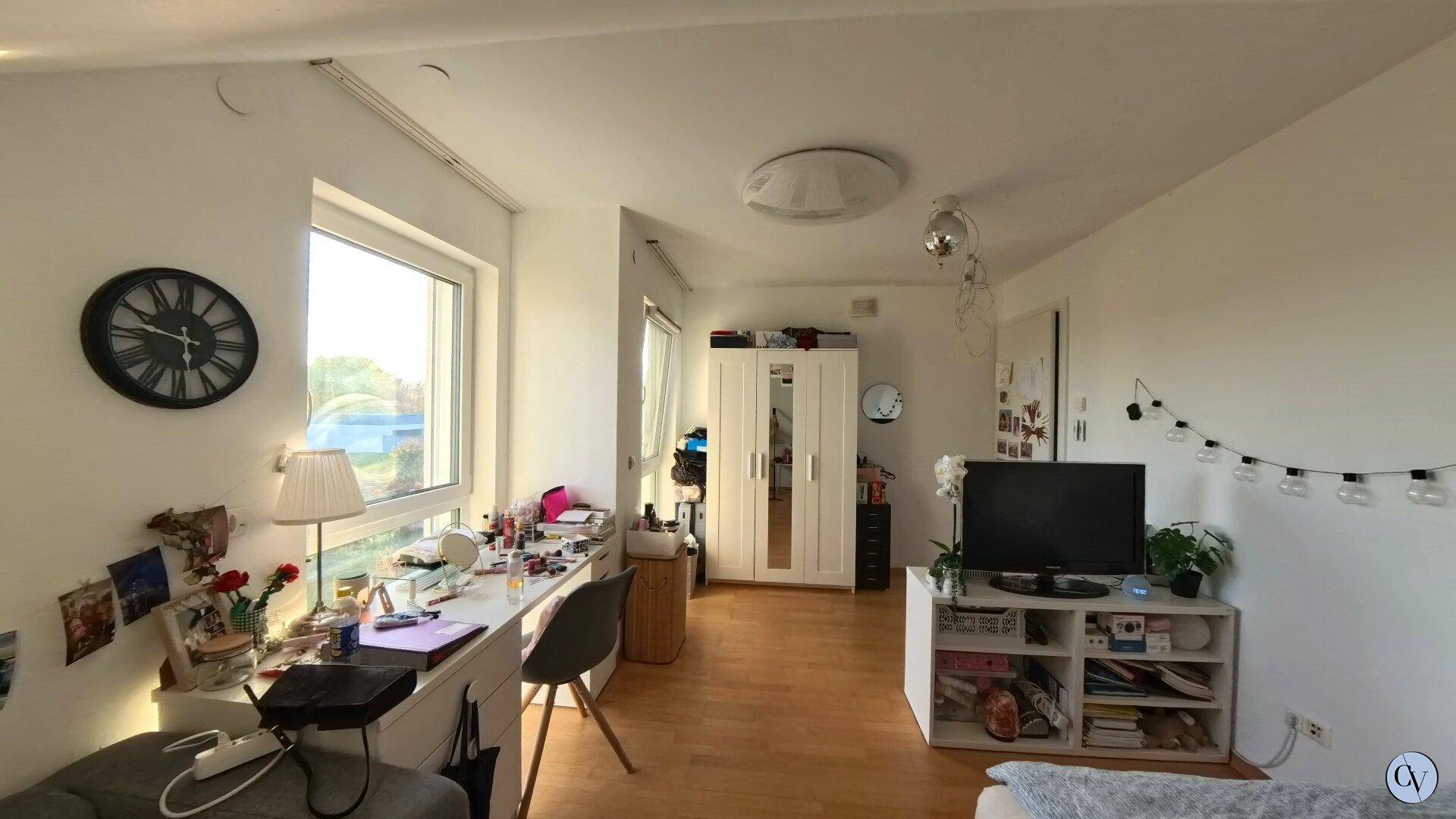 Helles Schlafzimmer mit Schreibtisch, Kleiderschrank und Fenster mit Blick nach draußen.