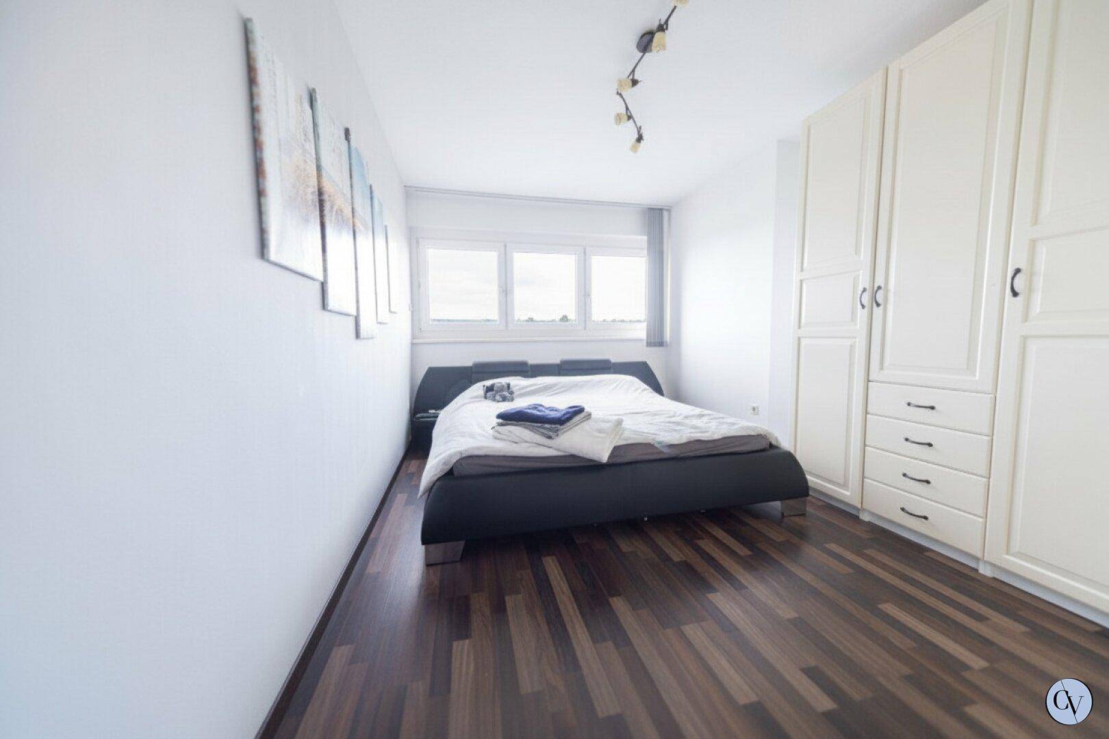 Schlafzimmer mit dunklem Laminatboden, weißem Kleiderschrank und großen Fenstern.
