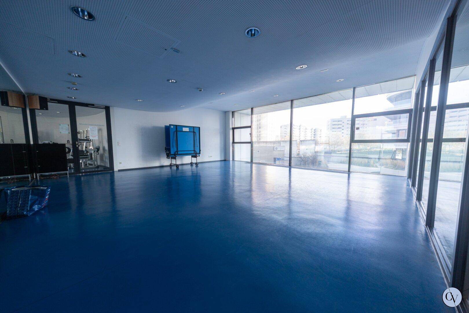 Großer Gemeinschaftsraum mit blauem Boden, großen Fenstern und Tischtennisplatte.
