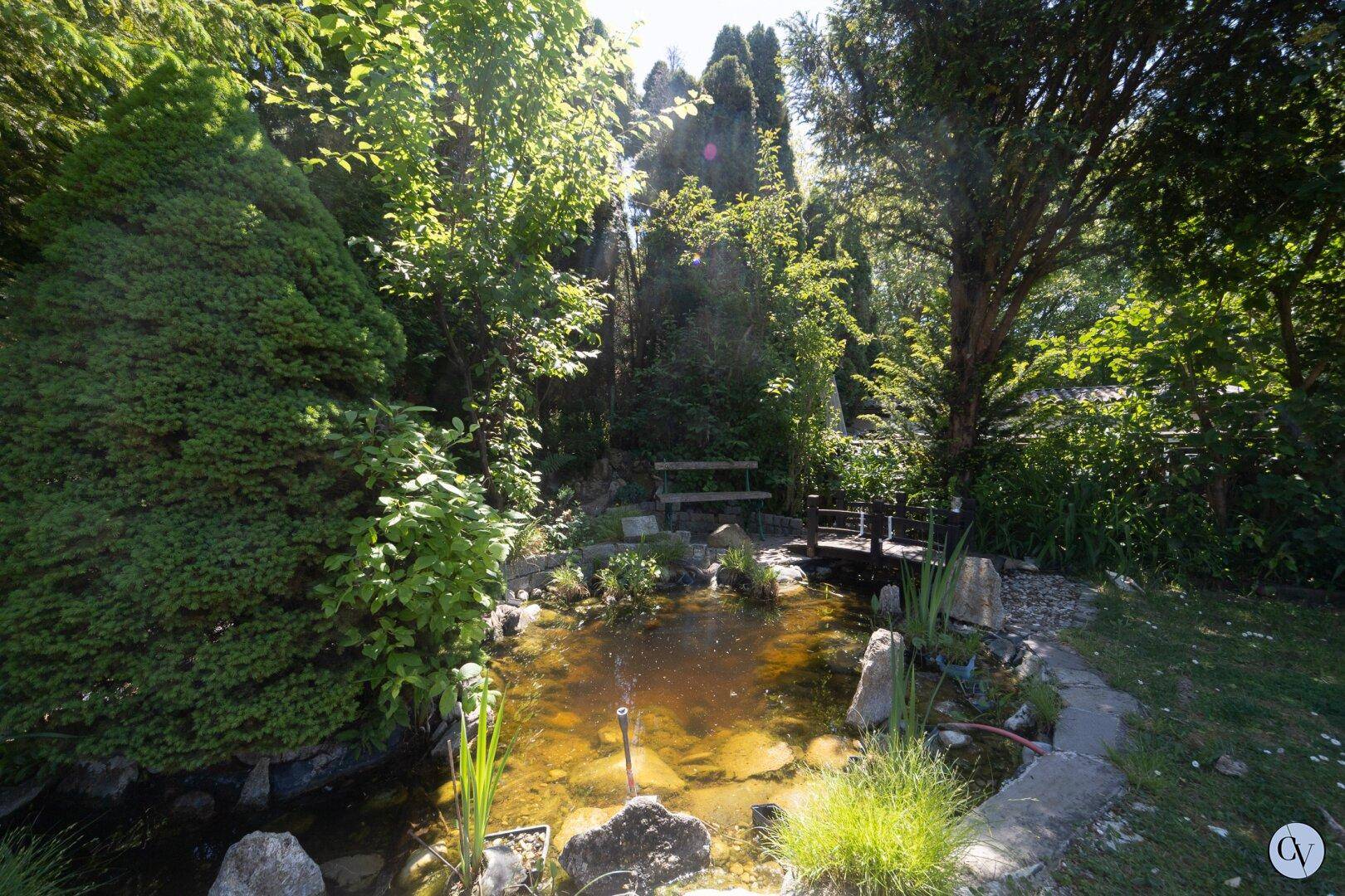 Idyllischer Gartenteich umgeben von üppigem Grün, Bäumen und einer kleinen Holzbrücke.