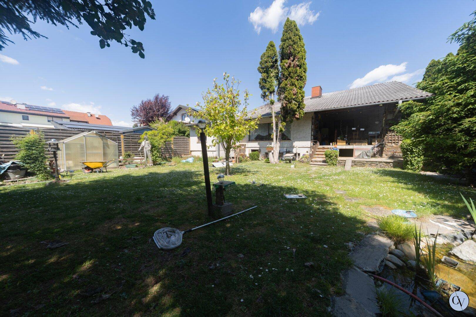 Großer Garten mit Rasenfläche, Bäumen, Gewächshaus und überdachter Terrasse am Haus.