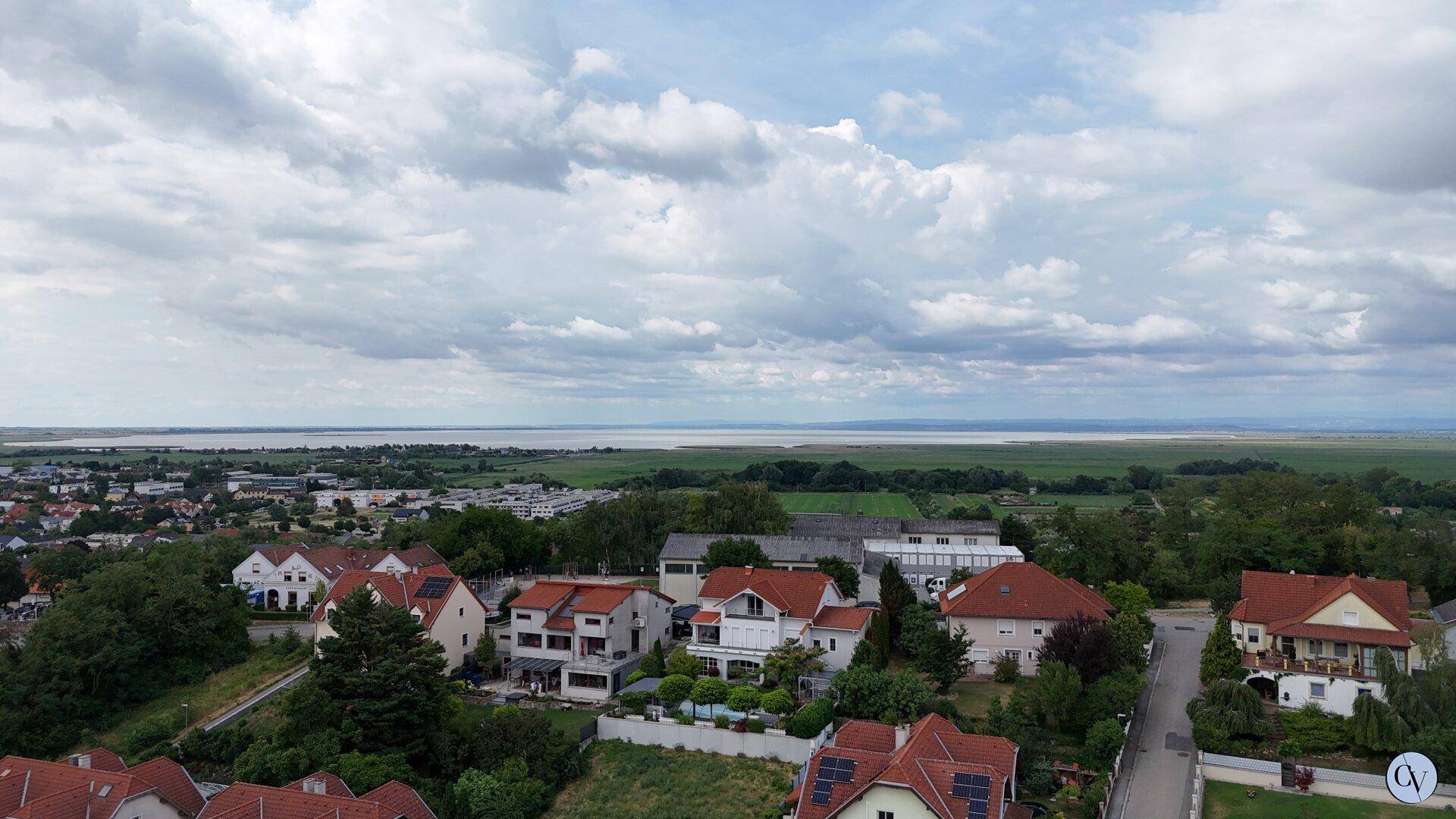 Panoramablick auf eine Siedlung mit Häusern, grünen Feldern und einem See in der Ferne unter bewölktem Himmel.