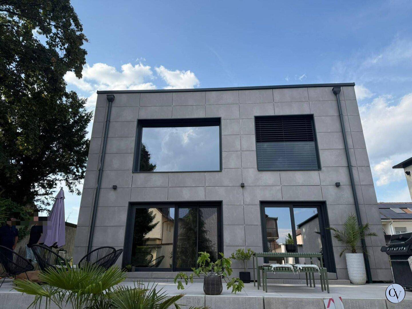 Moderne Hausfassade mit großen Fenstern und einer möblierten Terrasse unter blauem Himmel.