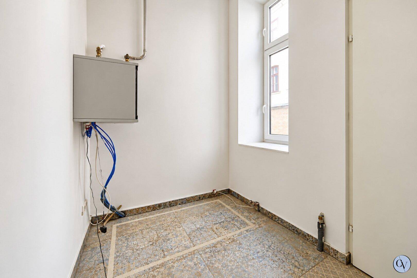 Kleiner Raum mit weißen Wänden, Fliesenboden, einem Fenster und sichtbaren Installationen.