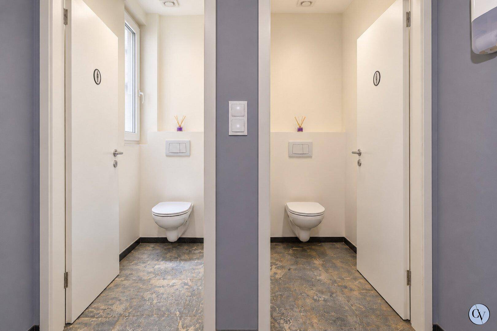 Zwei separate Toilettenräume mit modernen Wand-WCs und Fliesenboden, getrennt durch eine Wand.