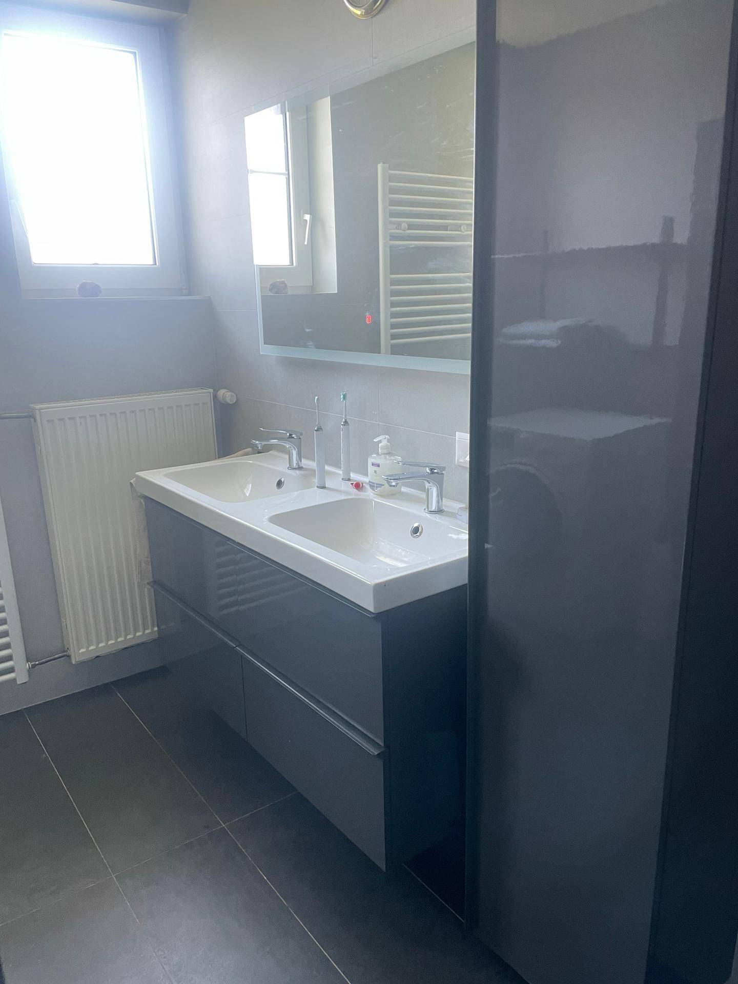 Helles Badezimmer mit Doppelwaschbecken, Spiegelschrank, Fenster und Handtuchheizkörper.