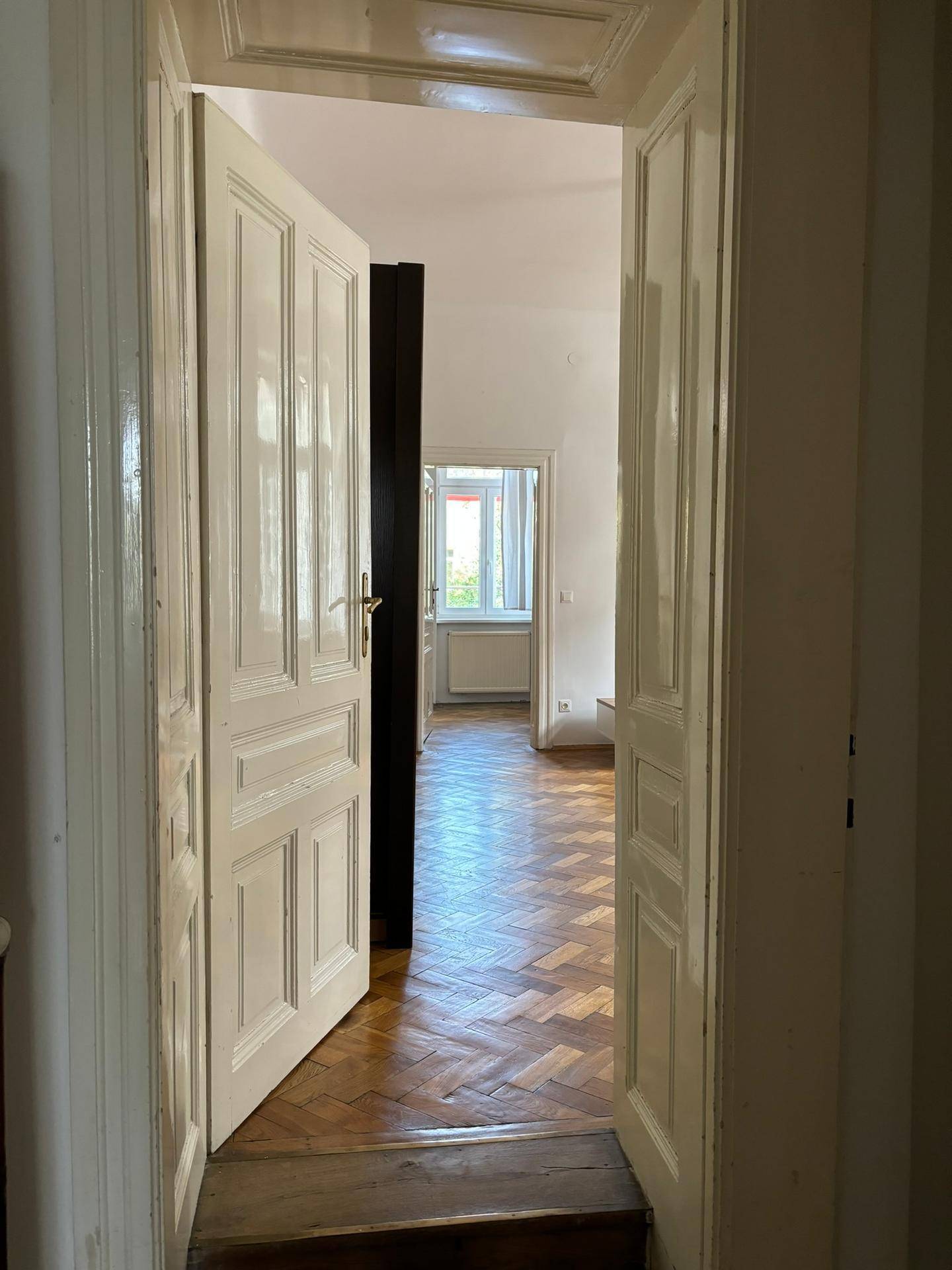 Blick durch eine offene Tür in einen weiteren Raum mit klassischem Fischgrätparkett und viel Tageslicht.