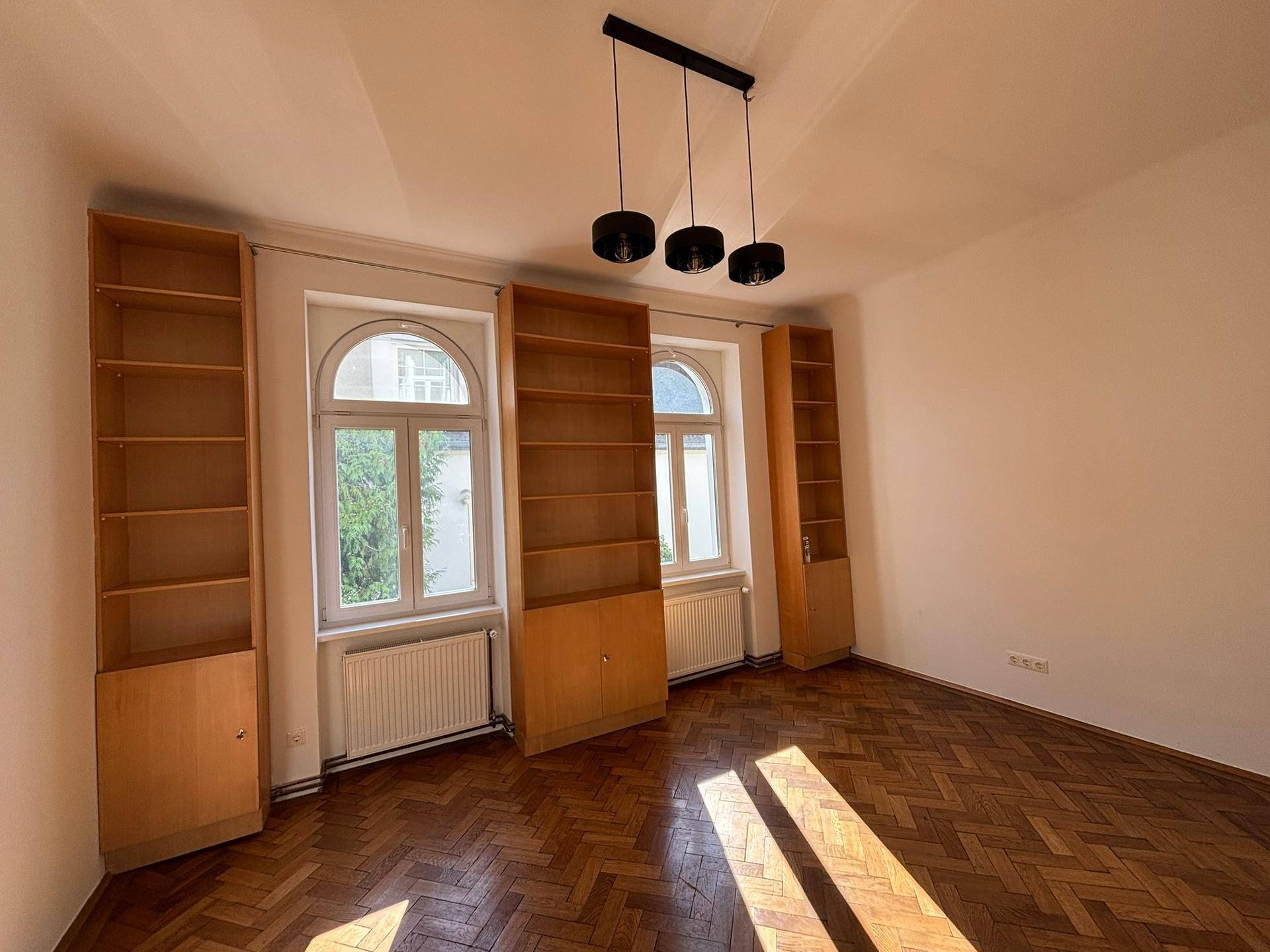 Geräumiges Wohnzimmer mit hohen Fenstern, integrierten Holzregalen und sonnigem Fischgrätparkettboden.